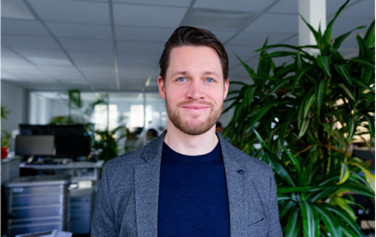 Ein lächelnder Mann steht in einem modernen Büro mit Pflanzen im Hintergrund.