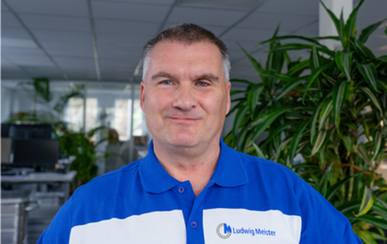 Ein Mann in einer blau-weißen Uniform steht lächelnd in einem Büro mit Pflanzen im Hintergrund.