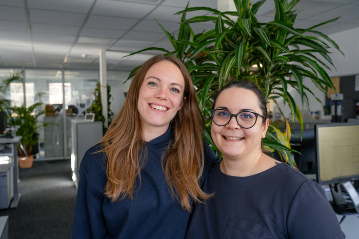 Zwei Frauen lächeln in einem modernen Büro mit Pflanzen im Hintergrund in die Kamera.