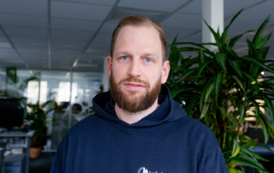 Ein Mann mit Bart und dunkelblauem Hoodie steht in einem Büro vor einer Pflanze.