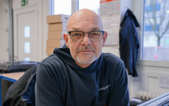 Ein älterer Mann mit Brille und grauem Bart sitzt in einem Büro, umgeben von Kartons und einem Kalender an der Wand.