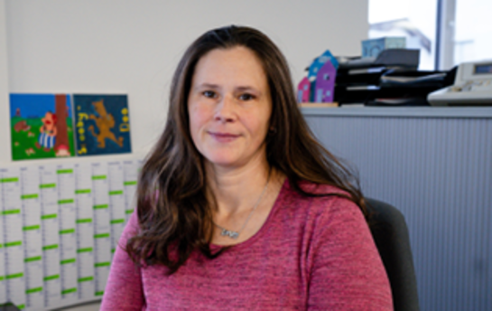 Eine Frau in einem roten Pullover sitzt in einem Büro mit einem Kalender und bunten Bildern an der Wand im Hintergrund.