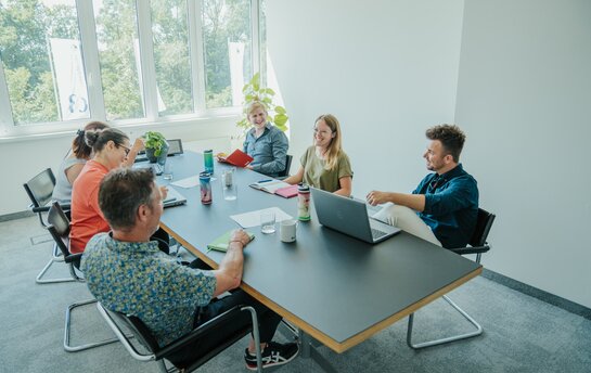 Eine Gruppe von sechs Personen sitzt in einem modernen Konferenzraum zusammen und unterhält sich angeregt.