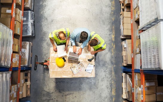 Drei Personen in Warnwesten stehen an einem Tisch im Lager und betrachten einen Laptop.