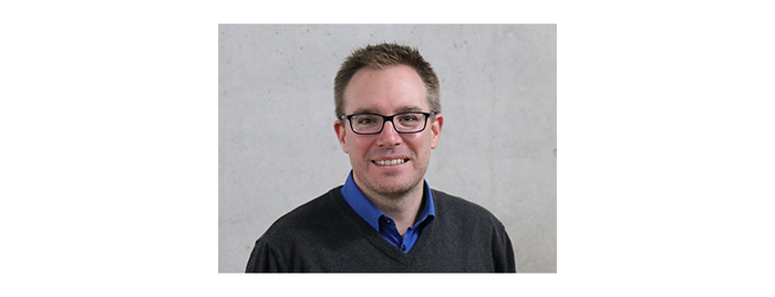 Ein Mann mit Brille und kurzem Haar lächelt in die Kamera, vor einem neutralen Hintergrund.