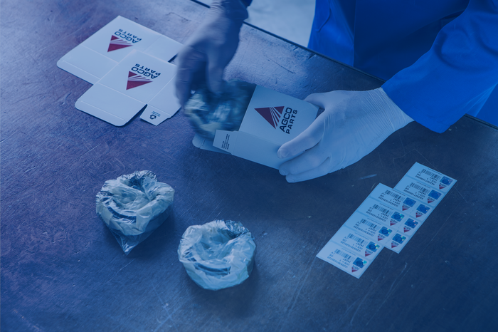 Eine Person verpackt ein Produkt in einer AGCO Parts-Box auf einem Tisch, zusammen mit etikettierten Aufklebern und weiteren Teilen.