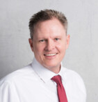 Ein Mann in einem weißen Hemd und roter Krawatte lächelt vor einem neutralen Hintergrund.