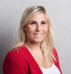 Eine lächelnde Frau mit blonden Haaren trägt einen roten Blazer vor einem grauen Hintergrund.