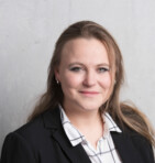 Eine Frau in einem schwarzen Blazer und kariertem Hemd lächelt vor einem grauen Hintergrund.