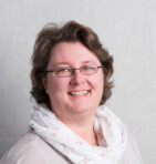 Eine lächelnde Frau mit Brille und hellem Schal vor einem neutralen Hintergrund.