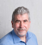 Ein Mann mit grauem Haar und einem blauen Hemd lächelt vor einem grauen Hintergrund.