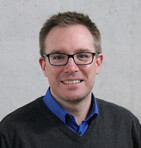 Ein lächelnder Mann mit Brille und blauem Hemd posiert vor einer grauen Wand.