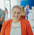 Eine Frau in einem orangefarbenen Blazer lächelt in die Kamera, während im Hintergrund Personen bei einem Empfang interagieren.