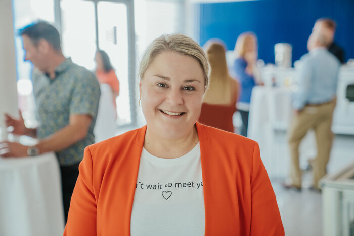 Eine Frau in einem orangefarbenen Blazer lächelt in die Kamera, während im Hintergrund Personen bei einem Empfang interagieren.