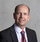 Ein Mann in einem Anzug mit Krawatte steht vor einer grauen Wand und lächelt.