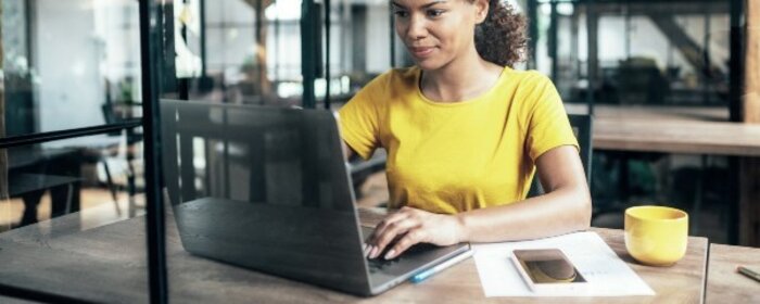 Eine Person in einem gelben T-Shirt arbeitet an einem Laptop in einem modernen Büro mit einer gelben Tasse neben sich.