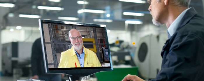 Ein Mann in Arbeitskleidung sitzt vor einem Monitor und nimmt an einer Videokonferenz mit einem anderen Mann in gelber Jacke teil, während er sich in einer Fabrikumgebung befindet.