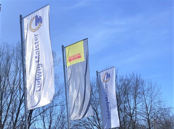 Drei Fahnen von Ludwig Meister und Sandvik flattern vor einem blauen Himmel im Wind.