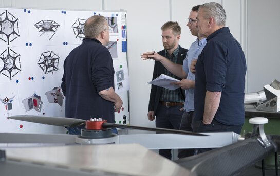 Vier Männer stehen vor einem Whiteboard mit technischen Zeichnungen und diskutieren.