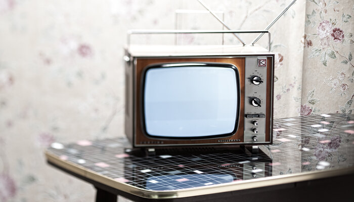 Ein alter Röhrenfernseher mit Holzrahmen steht auf einem schwarz-weiß-karierten Tisch vor einer geblümten Tapete.