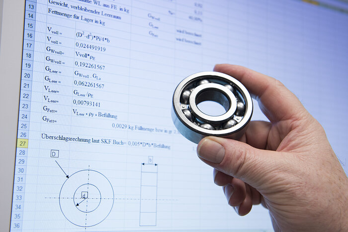 Eine Hand hält ein Kugellager vor einem Computerbildschirm, der Zeichnungen und Berechnungen anzeigt.