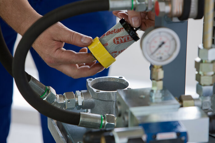 Eine Person ersetzt einen Hydraulikfilter in einem technischen Gerät mit verschiedenen Anschlüssen und einem Manometer.